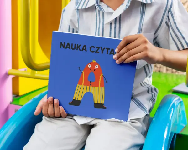 Chłopiec trzyma książkę "Nauka czytania dla 6 latka" z zabawną literką "A" na okładce.