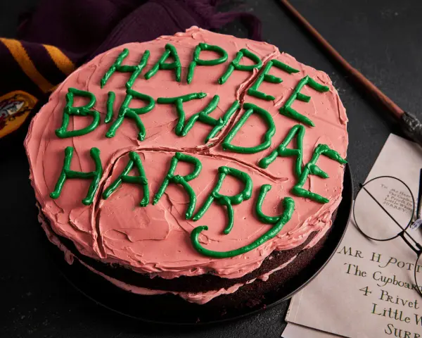 Ein Harry Potter Kuchen mit rosa Zuckerguss und grüner Schrift "Happy Birthday Harry". Daneben liegen ein Schal, eine Brille und ein Brief.