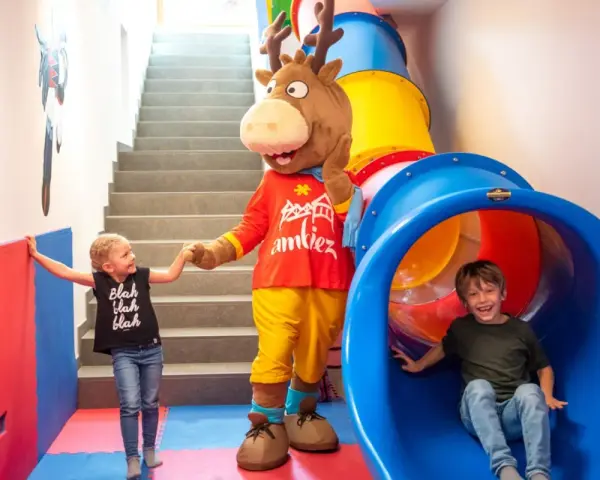 Mascot and two children in a colorful indoor playground with a slide and stairs.