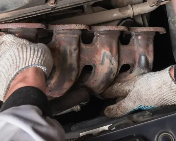 Mechanic's gloved hands work on a rusty exhaust manifold, possibly to fix an engine ticking noise.