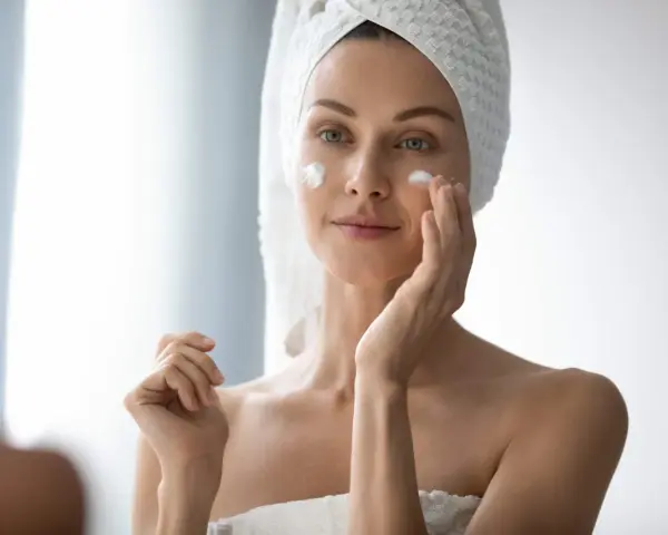 Femme appliquant de la crème sur son visage pour lutter contre la xerose cutanée. Elle a une serviette sur la tête.