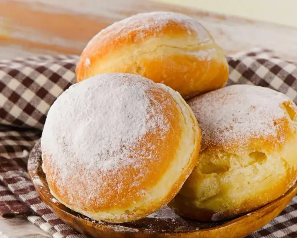 Trzy puszyste pączki, dwa posypane cukrem pudrem, leżą na drewnianej misce. Idealne na tłusty czwartek, te fit pączki kuszą apetycznym wyglądem.