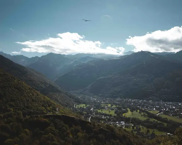 Vue aérienne d'une vallée verdoyante avec un village niché, entouré de montagnes majestueuses. Un planeur plane dans le ciel bleu, rappelant les paysages de Toulouse et ses environs.