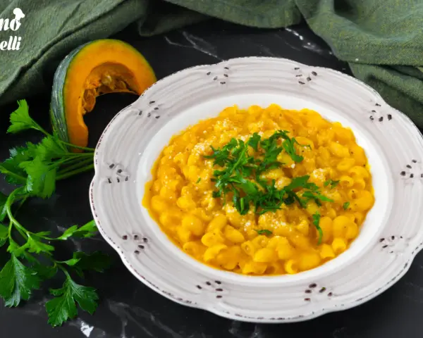 Piatto di pasta e zucca alla napoletana, guarnito con prezzemolo fresco e parmigiano. Accanto, una fetta di zucca e un mazzetto di prezzemolo.