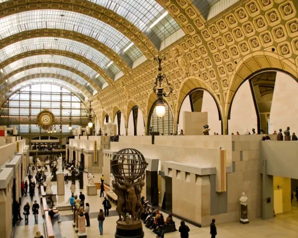 Interior del museo d'Orsay, con su impresionante techo de cristal y arcos dorados. Visitantes pasean entre esculturas y obras de arte.