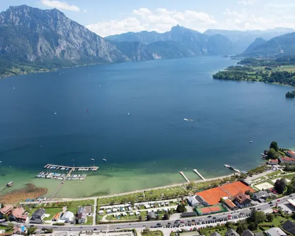 Idyllischer **Camping Traunsee** mit Blick auf den See und die Berge. Boote liegen im Hafen, Tennisplätze und Wohnwagen sind zu sehen.