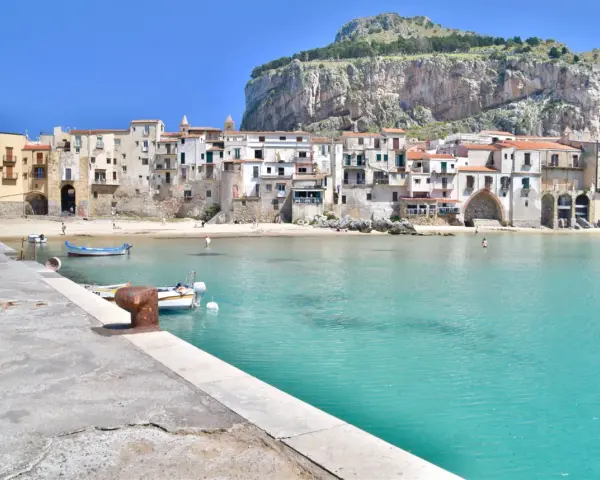 Coastal town with buildings nestled against a cliff, turquoise water, and boats. Perfect for a 10 days in Sicily adventure.