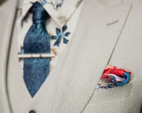 Elegancki mężczyzna w jasnej marynarce, z niebieskim krawatem i spinką do krawata jak nosić. W kieszeni kolorowa poszetka.