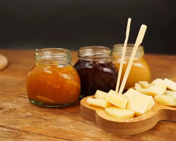 Tre vasetti di marmellata, uno arancione, uno scuro e uno giallo, accompagnano un tagliere di formaggio a cubetti, pronto per essere gustato.