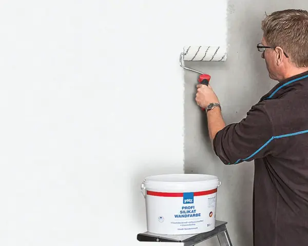 Mann streicht mit einer Farbrolle eine Wand weiß. Der Eimer mit der "Profi Silikat Wandfarbe" steht daneben. Perfekt für das Streichen bei Auszug.