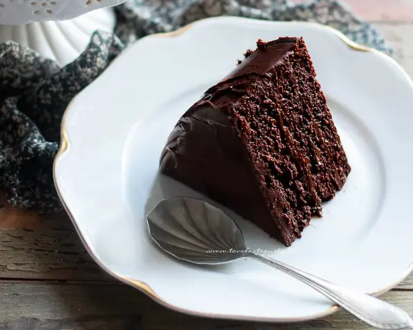 Una fetta di torta al cioccolato morbida e umida, ricoperta di glassa lucida, su un piatto bianco con un cucchiaino.