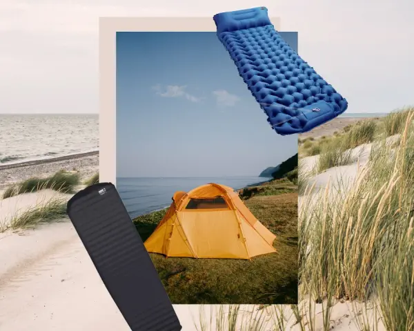 Zwei Isomatten, eine blaue aufblasbare und eine schwarze Schaumstoffmatte, liegen neben einem orangefarbenen Zelt am Strand.