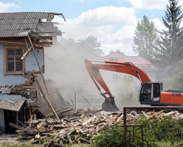 Koparka burzy zniszczony budynek, unosząc tumany kurzu. W tle inne domy i drzewa. Rozbiórka budynku formalności w toku.