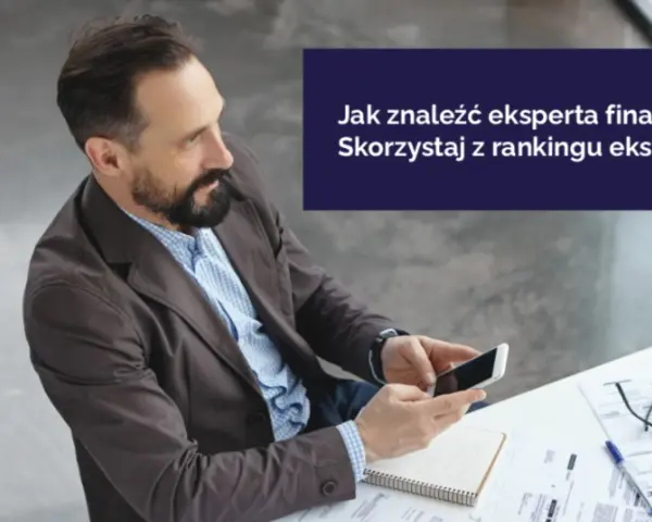 Mężczyzna z brodą trzyma smartfon, siedząc przy biurku z dokumentami.