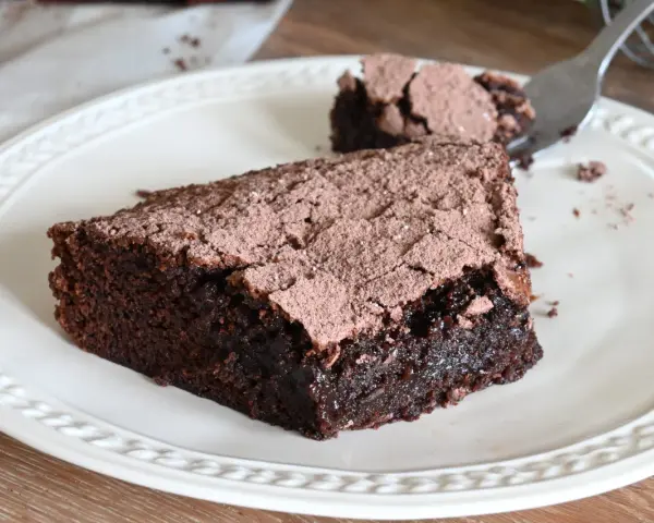 Una fetta di torta al cioccolato fondente, cosparsa di cacao, su un piatto bianco. Un pezzetto è stato staccato con una forchetta.