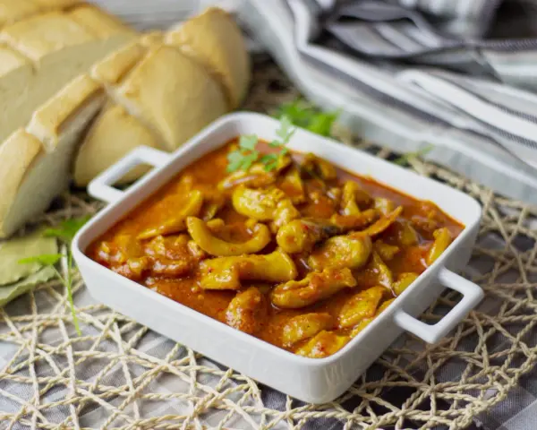 Delicioso plato de sepia en salsa, servido en cazuela blanca, acompañado de pan rústico y hojas de laurel.