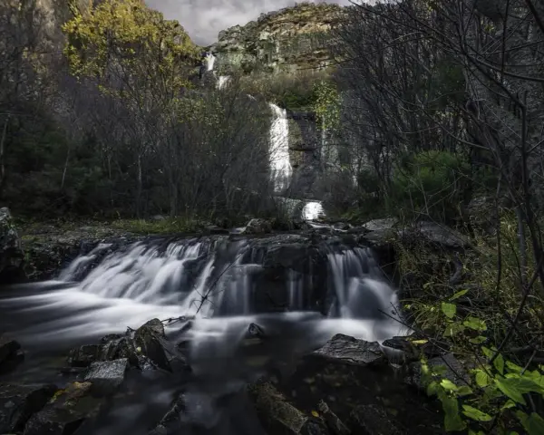 Cascada en el **río Bornova**, con aguas sedosas cayendo entre rocas y vegetación otoñal, bajo un cielo nublado.