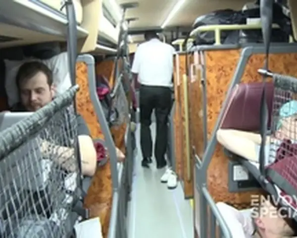 Intérieur d'un bus avec des couchettes. Un homme en blanc marche dans l'allée, tandis que des passagers se reposent dans leurs couchettes.