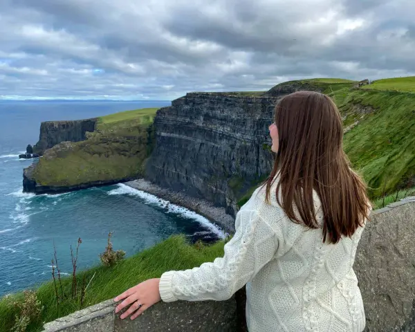 A woman in a cream cable-knit sweater gazes at the dramatic Cliffs of Moher, a breathtaking highlight of many tours of Ireland.
