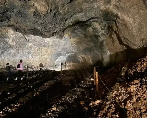 Wnętrze jaskini ciemnej, z kamienistym dnem i skalnymi ścianami. W oddali widać dwie osoby zwiedzające.