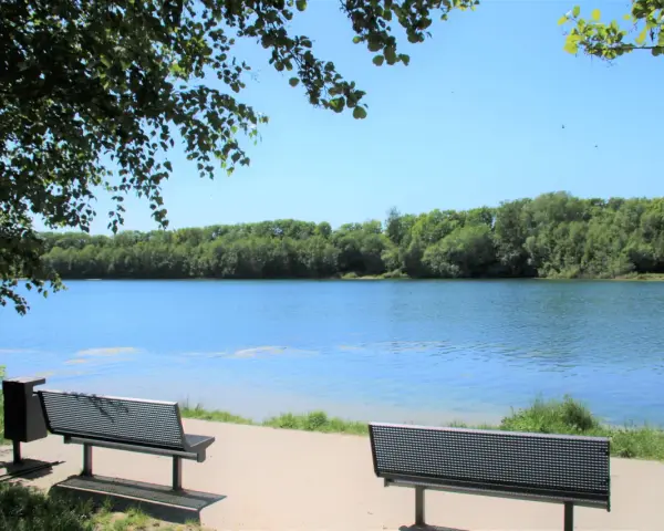 Zwei Bänke laden zum Verweilen am Ufer des halener Badesees ein. Bäume säumen das klare Wasser unter blauem Himmel.