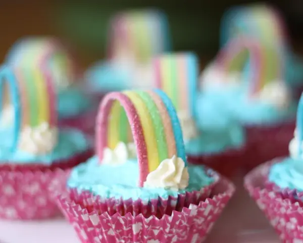 Sju cupcakes med ljusblå regnbågs frosting och godisregnbågar. Rosa muffinsformar med mönster.
