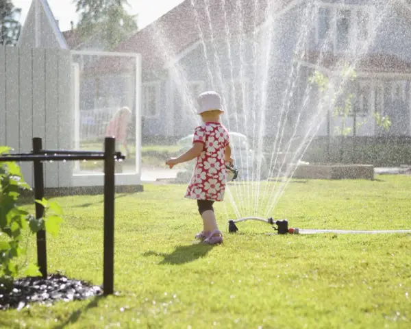 Lekfull **tipspromenad barn** i sommarregn från en vattenspridare. Ett litet barn i blommig klänning njuter av vattnet på gräsmattan.