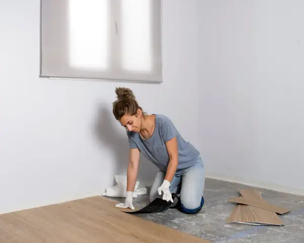 Mujer con guantes instalando suelo vinílico, colocando una tabla de madera clara sobre el suelo de hormigón.