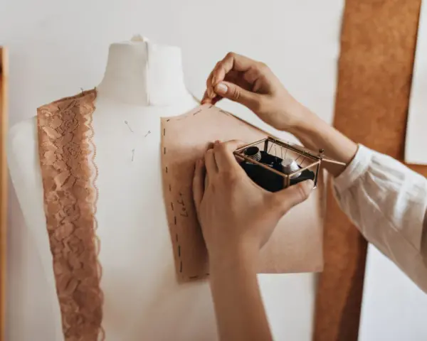 Mani che lavorano su un manichino, creando un modello di cartone. Un corso di modellistica in pieno svolgimento, con pizzi e accessori da cucito a portata di mano.
