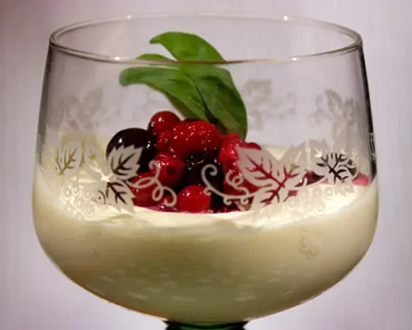 En elegant glas med vit chokladmousse, toppad med färska bär och ett grönt blad. Glaset har en vacker dekor av löv.
