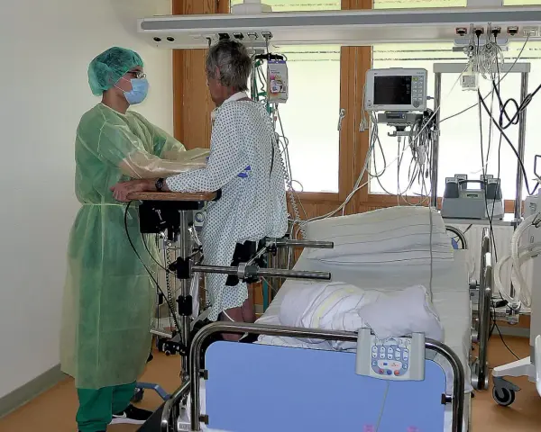 Pflegekraft unterstützt Patientin bei der Reha nach Intensivstation. Sie üben das Aufstehen.