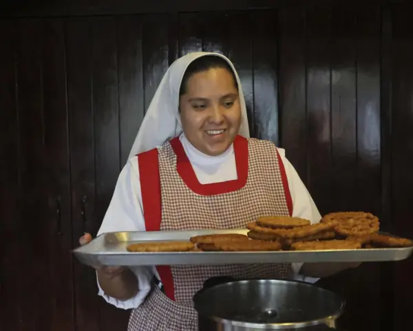 A smiling nun in a white habit and checkered apron holds a tray of fried pastries, perhaps answering the question: what do nuns eat for breakfast?