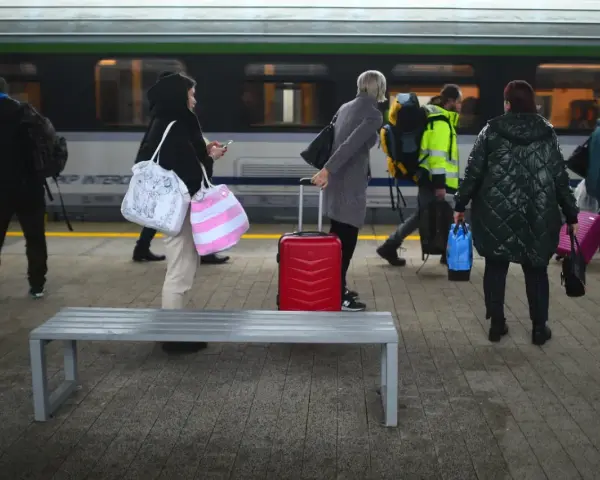 Ludzie z bagażami czekają na peronie PKP Intercity. Czerwona walizka stoi obok ławki.