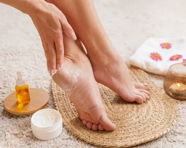 Application d'une crème hydratante sur la peau sous les pieds pour un soin doux. Une bougie parfumée et une serviette décorée complètent ce moment de détente.