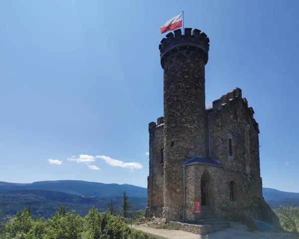 Kamienna wieża z polską flagą, część zamku księcia Henryka, góruje nad malowniczym krajobrazem gór i lasów.