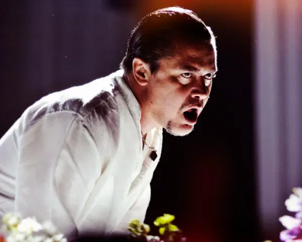 Mike Patton heute, mit weit aufgerissenem Mund und intensivem Blick, trägt ein weißes Hemd. Blumen im Vordergrund.