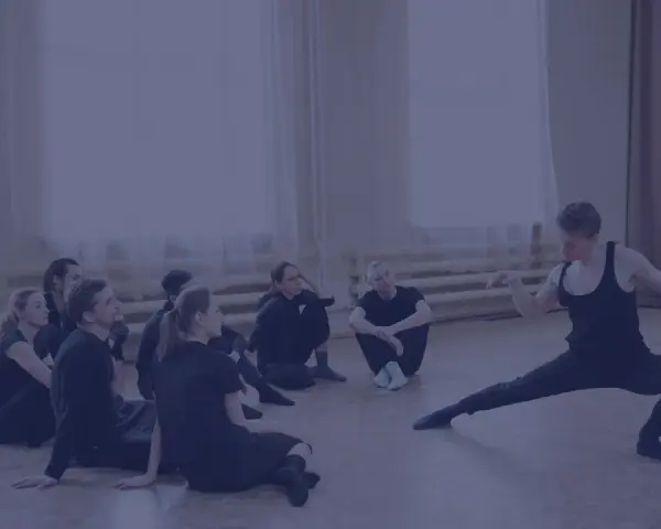 Choreograf demonstruje ruchy grupie tancerzy siedzących na podłodze.