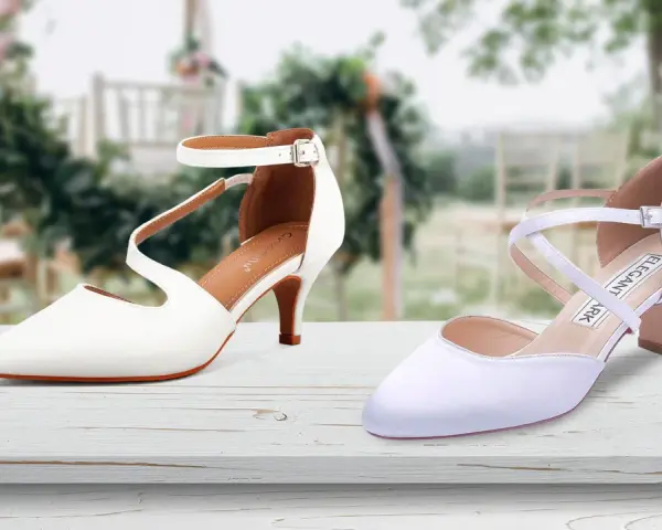 Due eleganti scarpe da sposa bianche, una con cinturino alla caviglia, l'altra con incroci sul collo del piede, su un tavolo di legno con sfondo sfocato di un matrimonio.