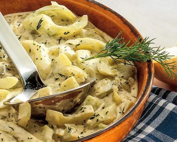 Cremiges Schmorgurken Rezept mit Dill in einer Tonschale, daneben eine Zwiebel und ein Löffel.
