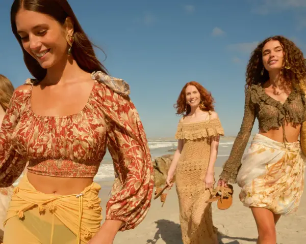 Quattro donne con abiti in stile boho passeggiano su una spiaggia sabbiosa con il mare sullo sfondo.