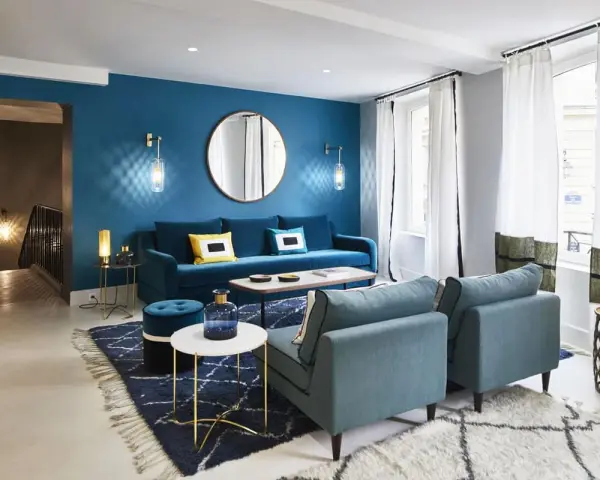 Salon moderne avec mur bleu canard, canapé velours, fauteuils et tapis assortis. Le jaune vif des coussins dynamise l'ensemble.