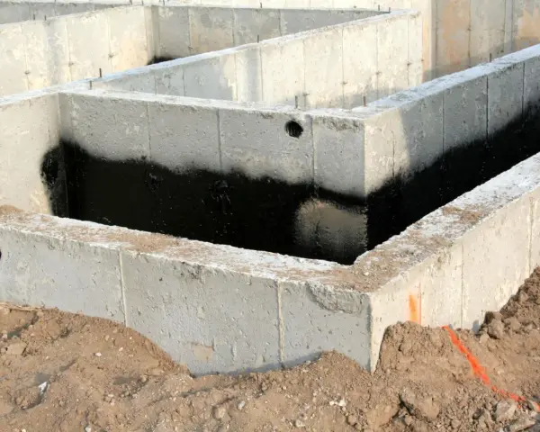Surowe ściany fundamentowe z betonu, częściowo pokryte czarną izolacją przeciwwilgociową, otoczone ziemią.