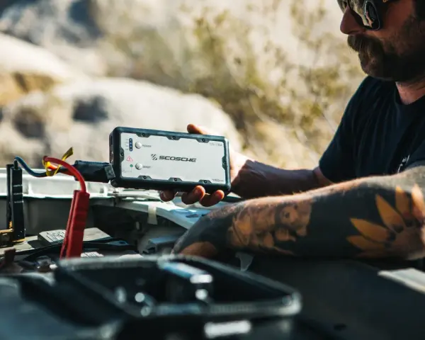 Man with tattoos demonstrates how to charge a jump starter by connecting it to a car battery in a desert landscape.