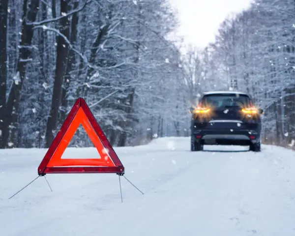 Warnblinkleuchten und Warndreieck auf verschneiter Straße. Ein Auto steht im Hintergrund. Ein wichtiger Hinweis für den auto wintercheck.