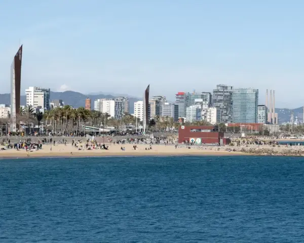 Ludzie odpoczywają na plaży, a w tle widać nowoczesne budynki Barcelony. To miasto leży nad Morzem Śródziemnym.