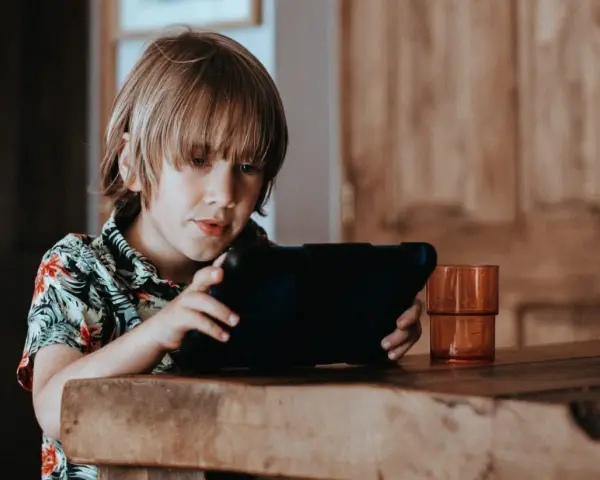 Chłopiec w hawajskiej koszuli ogląda krótkie filmy na tablecie, siedząc przy drewnianym stole.