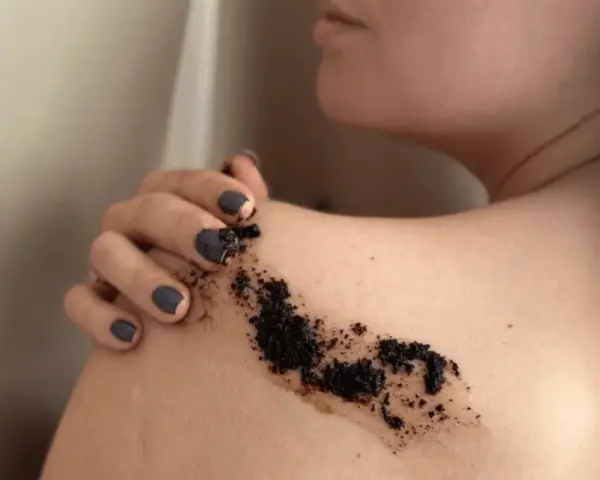 Dłoń z ciemnym lakierem nakłada peeling kawowy na ramię. Efekty po miesiącu regularnego stosowania są widoczne.