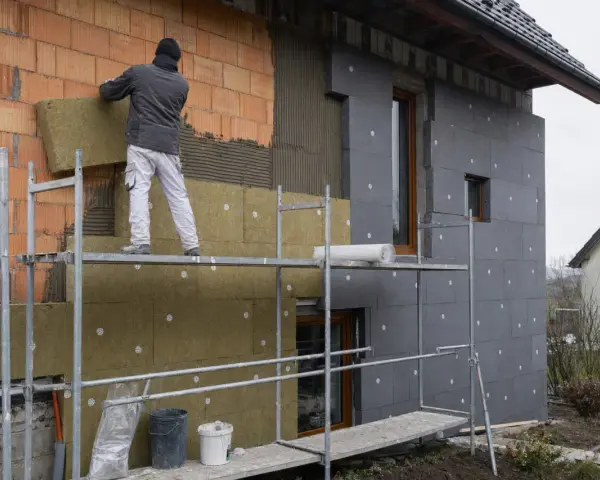 Pracownik na rusztowaniu montuje wełnę mineralną na ścianie domu. Brak cokołu na elewacji, trwa ocieplanie.