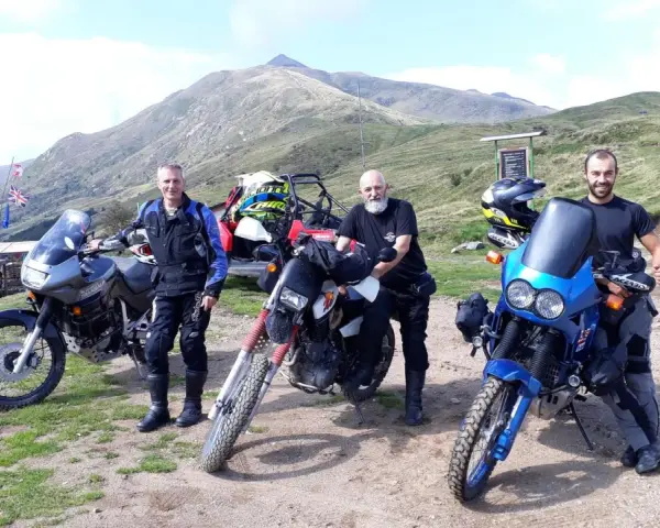 Tre motociclisti con moto da enduro posano in cima al passo San Lucio, con montagne sullo sfondo.