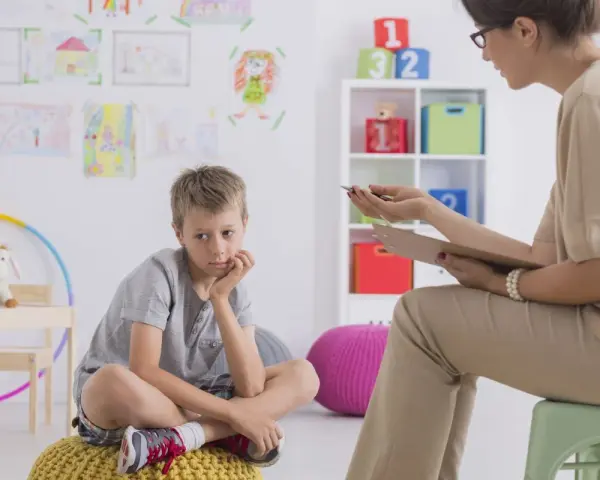 Chłopiec siedzi na poduszce, zastanawiając się, jakie pytania zadaje psycholog dziecku. W tle rysunki dzieci i kolorowe zabawki.